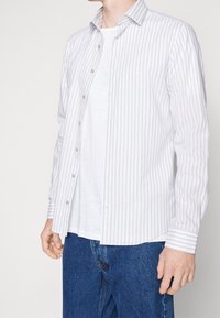 Pruhovaná košile na knoflíky v bílé a světle šedé barvě, dlouhé rukávy, s límečkem a knoflíkovou légou, nošená přes bílý tričko a modré džíny.