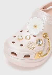 Sabot rose clair avec des trous de ventilation décoré de fleurs blanches, de breloques en perle et cristal, et d'un accessoire chaîne dorée.