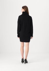 Crna pletena haljina s turtleneck izrezom, dugih rukava i opuštenog kroja, koja doseže do sredine bedra, u kombinaciji s crnim stiklama.