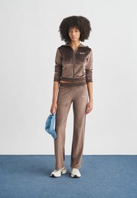 Survêtement en velours marron avec veste zippée et pantalon assorti, avec un petit logo. Le mannequin tient une casquette bleu clair et porte des baskets blanches.