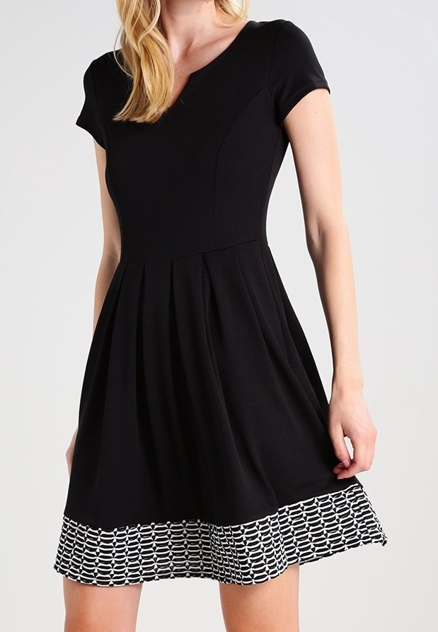 Femme portant une robe noire à manches courtes avec un col en V et un ourlet noir et blanc à motifs, debout devant un fond clair uni.