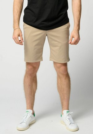 Uomo che indossa pantaloncini beige, scarpe da ginnastica bianche con accenti verdi e una maglietta nera, in piedi davanti a uno sfondo chiaro e uniforme.