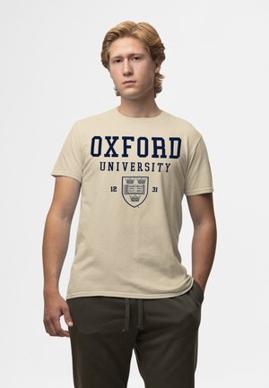 Giovane uomo con capelli chiari che indossa una maglietta beige dell'Università di Oxford e pantaloni scuri, in piedi con una mano in tasca davanti a uno sfondo semplice.