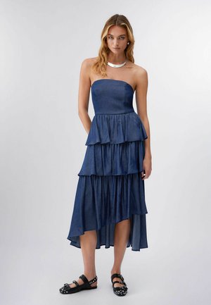 Donna che indossa un abito midi a balze blu navy senza spalline e scarpe basse nere con borchie, in piedi contro uno sfondo chiaro e semplice.