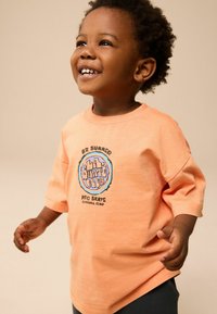 Oranje t-shirt met korte mouwen, voorzien van een ronde grafische logo in blauw en zwart met tekst. De stof lijkt zacht en heeft een casual pasvorm.