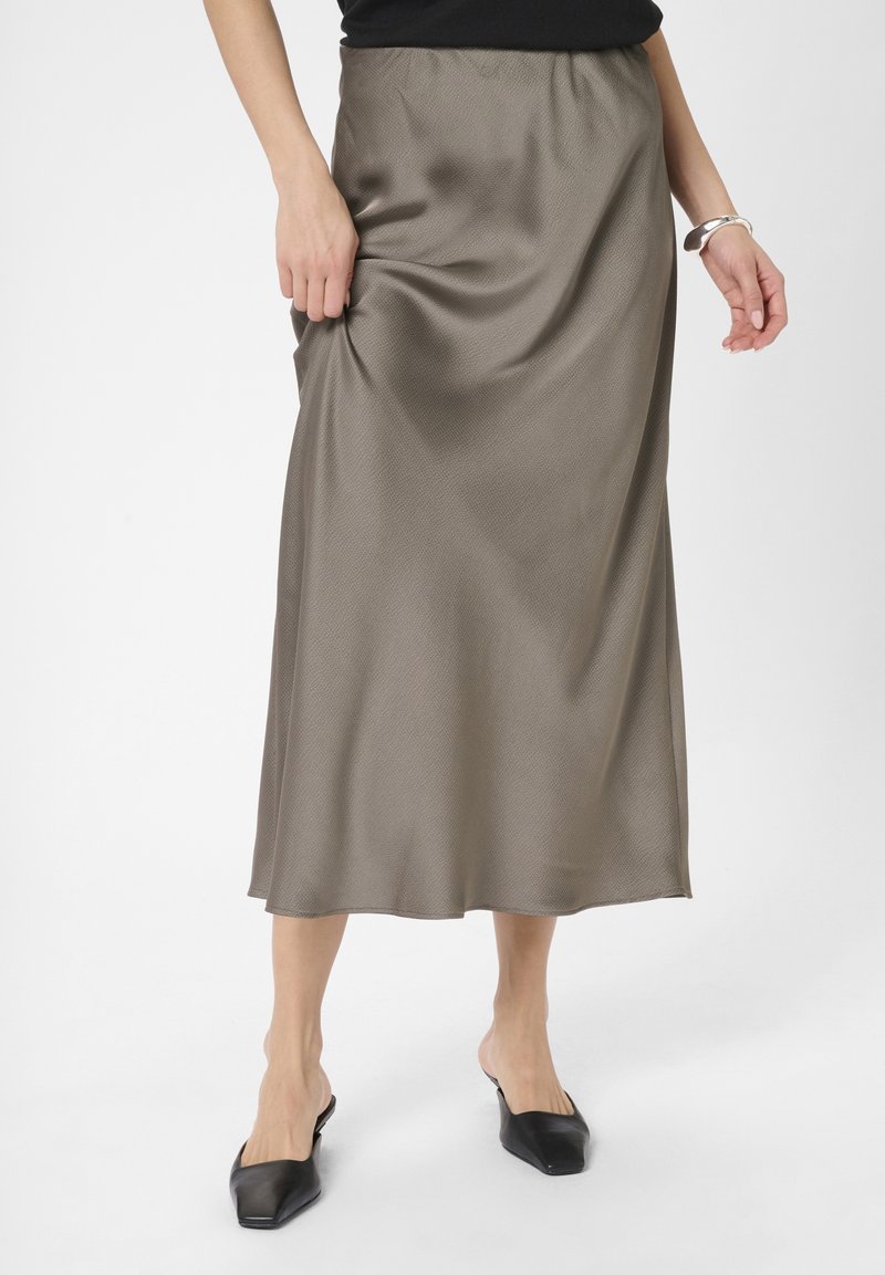 Femme portant une longue jupe en satin taupe, un haut noir sans manches, un bracelet argenté et des chaussures noires à bout pointu sans lacets, debout sur fond blanc.