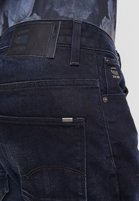 Mörkblå denimjeans med en slät textur, som har en svart läderlapp, silverfärgade detaljer och en signaturlogo på bakfickan.