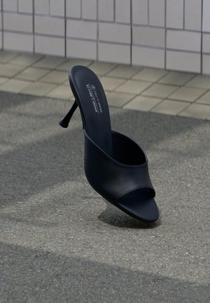 Must avatud kannaga king Mule-s stiilse kõrge kontsaga, asetatud asfaldile beeži ruudukujulise plaaditud seina lähedal, tallasisel tekstiga marki ja taimse materjali kohta.