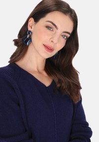 Pulover bleumarin tricotat cu decolteu în V, cu textură reiată și detaliu delicat de tricotaj cu cabluri. Cercei mari din acril albastru cu accente aurii.