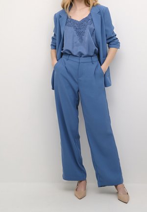 Vrouw draagt een blauw hemdje met kanten rand, bijpassende blauwe blazer met opgerolde mouwen, wijde broekspijpen en beige puntige hakken.