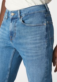 Jasnoniebieskie dżinsy o wysokim stanie, z delikatnym wyblaknięciem i pomarańczowymi przeszyciami. Zawierają zapięcie na guzik i zamek błyskawiczny oraz przednie kieszenie.