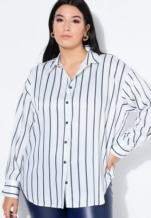 Frau mit langem dunklem Haar trägt ein weißes, knöpfbares Hemd mit vertikalen schwarzen Streifen und dunkle Hose, posiert mit einer Hand an der Hüfte vor weißem Hintergrund.