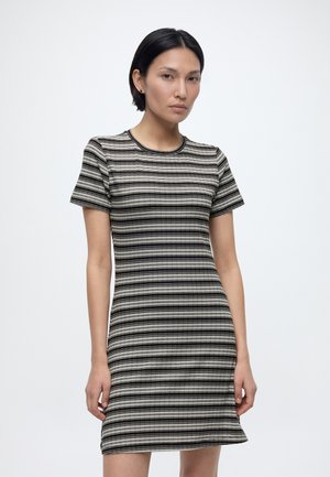 Žena s krátkymi čiernymi vlasmi, oblečená v krátkom páse pruhovaných čierno-bielo-šedých šatách, stojaca pred obyčajným bielym pozadím.