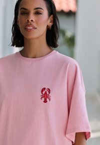 Lyserød t-shirt i bomuld med en afslappet pasform, med et rødt broderet hummelmotiv på venstre side af brystet. Korte ærmer.