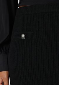 Fustă tricotată neagră cu o textură ribbed, un buzunar lateral și un nasture metalic de culoare argintie cu un detaliu în formă de "G". Prim-plan al țesăturii și designului.