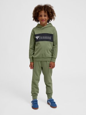 Dziecko o kręconych włosach, ubrane w oliwkowozielony kaptur hummel i dopasowane spodnie, stojące prosto w niebieskich sneakersach na jednolitym tle.