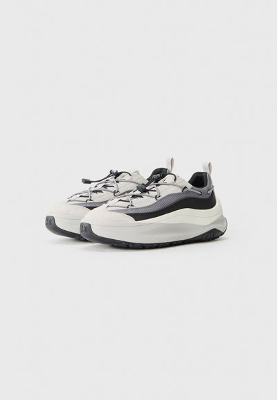 Zapatillas deportivas gris claro con ribete negro, parte superior de malla texturizada, suela de goma y cordones elásticos. Diseño curvado y talón acolchado.