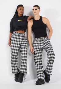 Svartvita rutiga byxor med elastisk midja; modellen är klädd med en kort svart topp och svarta sneakers, stående mot en vit bakgrund.