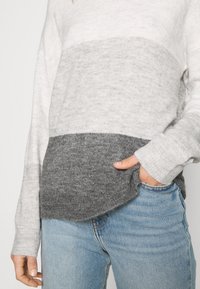 Pull gris et blanc à blocs de couleur avec un tricot texturé, présentant un ourlet arrondi et une coupe décontractée. Main reposant dans une poche de jeans bleus.
