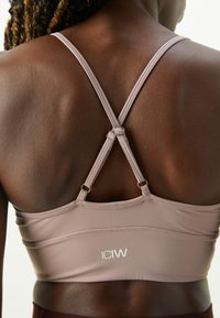 Reggiseno sportivo di un delicato colore malva con schiena aperta e design a incrocio delle spalline, caratterizzato da un tessuto morbido e un piccolo logo nella parte inferiore.