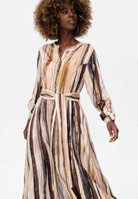 Lange, gestreepte jurk met een knoopsluiting aan de voorkant, beige, bruin en gouden tinten, en een strikceintuur. Zachte stof met lange mouwen.