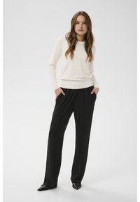 Krēmkrāsas džemperis ar ribotiem aprocēm, saskaņots ar melnām, kroku platām biksēm. Modelis stāv ar rokām kabatās, valkājot melnas, smailas kurpes.