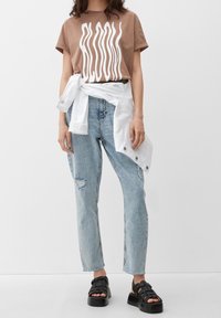 Hnědé tričko s bílým vlnitým motivem, světle modré roztrhané džíny, bílá košile zavázaná v pase, černé platformové sandály s pásky.