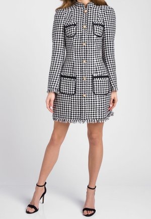 Donna che indossa un abito cappotto a manica lunga con motivo pied de poule nero e bianco, bottoni dorati e sandali neri con tacco alto.