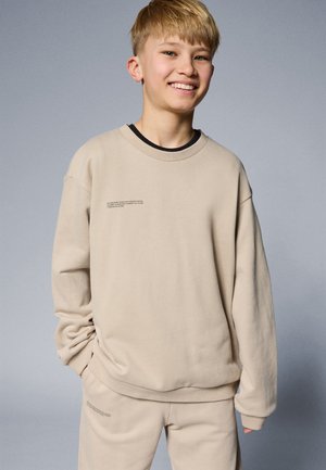 Beige sweatshirt with long sleeves, round neckline, and side pockets. Features small text detail on front. Soft material with a relaxed fit.