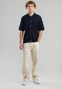 Tummansininen neule lyhythihainen paita napituksella, jossa on tekstuuri. Yhdistetty vaaleanbeigeihin housuihin ja valkoisiin lenkkareihin.