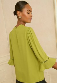 Zielona bluzka z bufkami, okrągłym dekoltem i tylnym rozcięciem w kształcie łezki. Posiada kontrastowe wykończenia u rękawów. Gładki, miękki materiał.