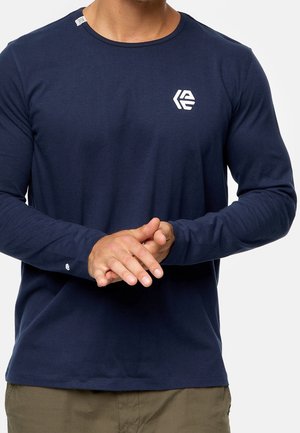 Mann trägt ein marineblaues Langarmshirt mit weißem Logo auf Brust und Ärmel, steht mit verschränkten Händen vorne.