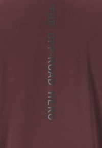 Bordeauxrotes T-Shirt mit vertikalem schwarzem Text, der "DER OFF-ROAD HELD" liest. Der Stoff hat eine dezente Struktur und eine taillierte Form.