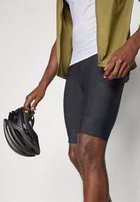 Schwarze Radhosen mit gepolstertem Einsatz, kombiniert mit einem weißen Netzshirt und einer olivgrünen Jacke mit Reißverschluss. Ein schwarzer Helm wird in einer Hand gehalten.
