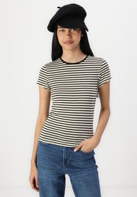 Stripet kortermet t-skjorte i krem og svart, med rund hals og tett passform, sammen med blå denimjeans og en svart hatt.