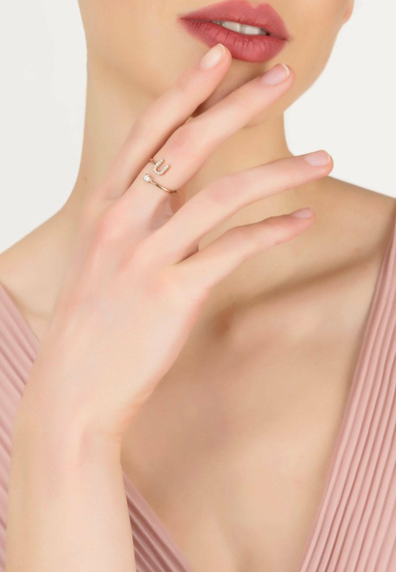 Gold rings on fingers; one features a diamond accent, the other has a ‘U’ design with small crystals. Hand rests near the chin.