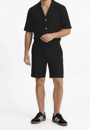 Mann trägt ein schwarzes kurzärmeliges Hemd mit Knöpfen, passende schwarze Shorts mit Kordelzug und schwarze Sneaker mit weißen Streifen.