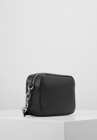 Černá kožená taška s texturovaným povrchem, čtvercového tvaru, na zip a se stříbrným řetízkem. Umístěná na bílé ploše.