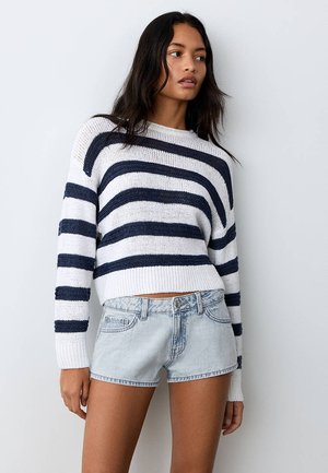 Kvinde iført en hvid og navystribet sweater og lyseblå denimshorts, der står mod en ensfarvet lys væg.