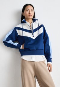 Pullover colorblock blu e azzurro con mezza zip, design a chevron prominente, tasche laterali e polsini a coste. Tessuto morbido.