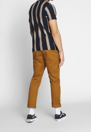 Homme portant une chemise à rayures verticales noires, blanches et marron, un pantalon marron retroussé, des chaussettes blanches et des baskets noires, debout devant un fond blanc.