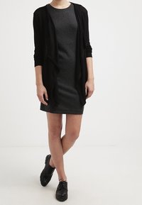 Cardigan negro sobre un vestido de canalé gris oscuro, hasta la rodilla, con un corte relajado. Combinado con zapatos negros con cordones, de diseño minimalista.