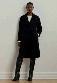 Γυναίκα που στέκεται φορώντας ένα μακρύ μαύρο παλτό με ζώνη, λευκό πουλόβερ και μαύρες δερμάτινες μπότες έως το γόνατο, μπροστά σε έναν απλό ανοιχτό τοίχο.