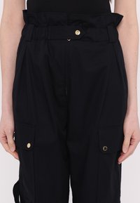 Pantalon cargo noir taille haute avec taille paperbag, détail ceinture boutonnée et poches à rabat sur les cuisses avant, porté avec un haut noir.