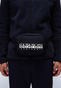 Marineblaue Fleecejacke mit einem halben Reißverschluss, kombiniert mit einer marineblauen Gürteltasche mit weißem Logo. Strukturierter, weicher Stoff mit verstellbarem Riemen.