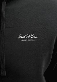 Μαύρο ύφασμα με λευκό κείμενο "Jack & Jones Archive Selection" κεντημένο στο στήθος και μαύρο κορδόνι στην αριστερή πλευρά.
