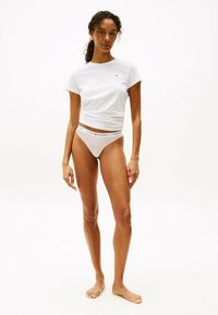 Witte katoenen t-shirt met opgerolde mouwen, gecombineerd met licht gestreepte bikiniondergoed met een merkband. Model poseert op blote voeten.