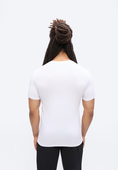 Άνδρας με μακριά πλεξούδες δεμένες σε κότσο, που φοράει εφαρμοστή λευκή κοντομάνικη μπλούζα και μαύρο παντελόνι, στέκεται με την πλάτη προς την κάμερα.