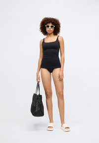 Fekete tankini felső négyzet alakú dekoltárral, fekete bikini alsóval párosítva. A modellen fekete hálós táska és krémszínű papucs található.
