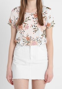 Mujer vestida con una blusa blanca de flores de manga corta metida dentro de una falda corta de mezclilla blanca con dobladillo desflecado, de pie contra un fondo liso.